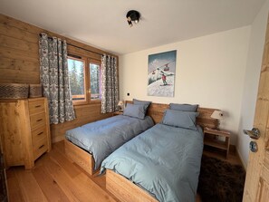 Ein Schlafzimmer mit zwei Betten, Holzwänden, einem Fenster mit Vorhängen, einem Gemälde und einer Holzkommode.