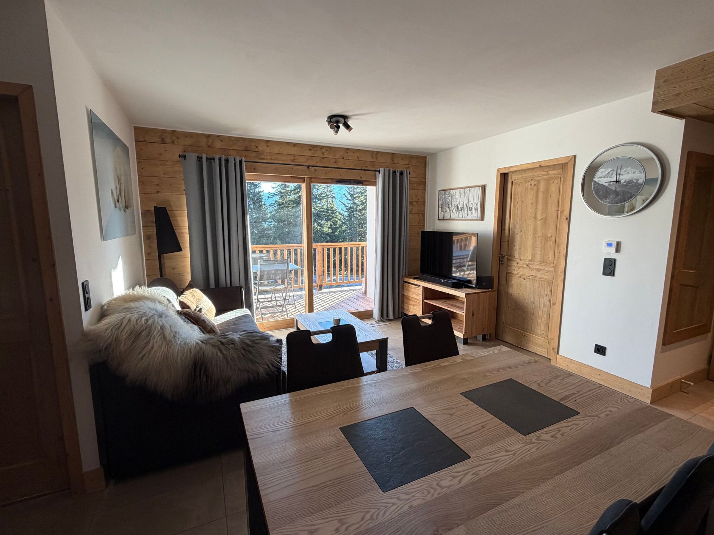 A modern living room with a wooden floor, a sofa, a dining table with chairs, a TV on a wooden stand, and a large window with curtains.