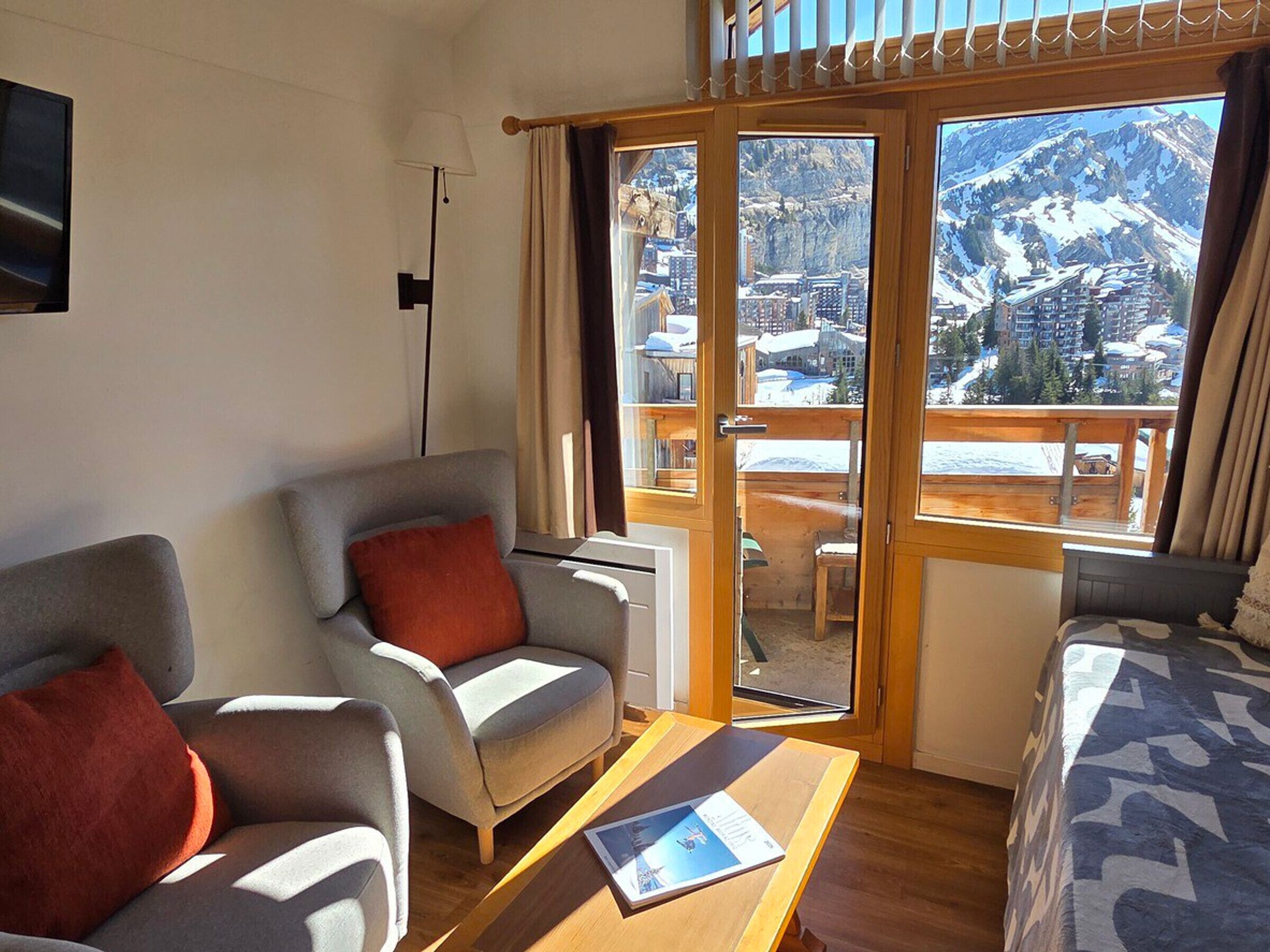 A hotel room with a sofa, a chair, a table, and a bed. There is a large window with a view of mountains and a city.