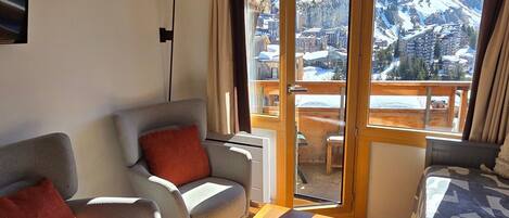 A hotel room with a sofa, a chair, a table, and a bed. There is a large window with a view of mountains and a city.