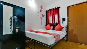 A single bed with red and white bedding, a wooden door, a black wardrobe, and a wall with a floral decoration. - Route 77 (Calangute)