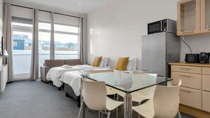 Habitación de hotel moderna con una mesa de comedor de cristal, dos camas, una kitchenette y vistas a la ciudad.