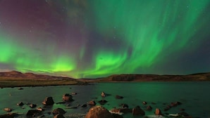 A natural landscape with a body of water, rocks, and a distant mountain range under a night sky with green aurora borealis.
