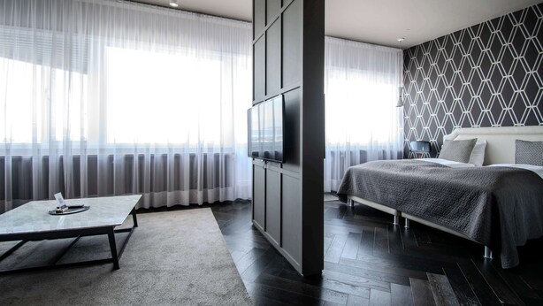 Ein modernes Schlafzimmer mit einem Bett, einem an der Wand befestigten Fernseher und einem großen Fenster mit durchscheinenden Vorhängen.