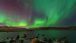 A natural landscape with a body of water, rocks, and a distant mountain range under a night sky with green aurora borealis. - Mani Apartments - Studio (Reykjavík)