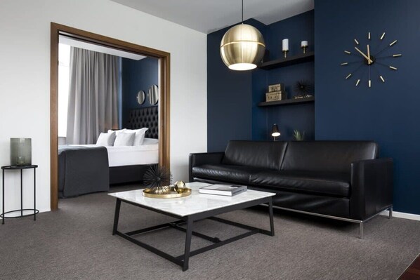 Una habitación de hotel moderna con un sofá de cuero negro, una mesa de centro blanca, un reloj de pared grande y una pared de acento azul oscuro.