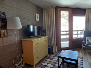 Una habitación con una televisión sobre un aparador de madera, una lámpara, una silla y vistas a través de una puerta.