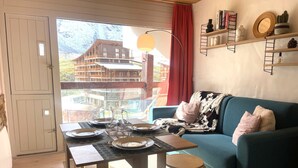 Un salon chaleureux avec un canapé bleu, une table à manger dressée pour quatre personnes et une vue sur un bâtiment en bois, visible à travers une porte vitrée.