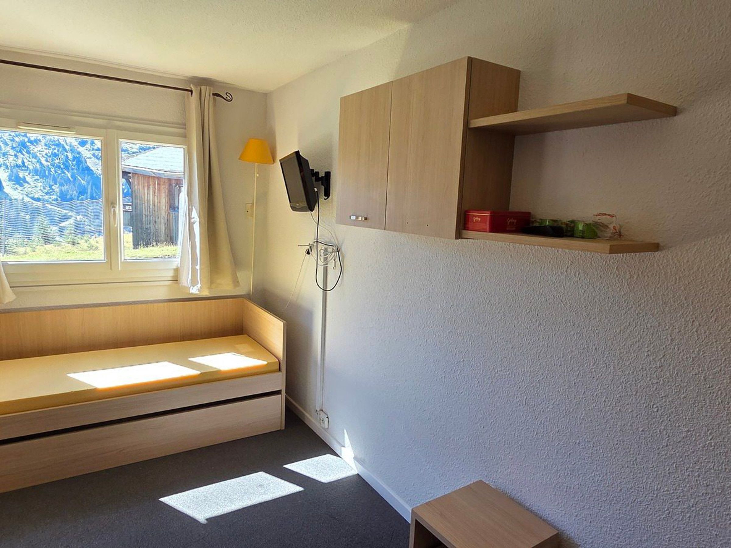 Une petite pièce agréable et chaleureuse, avec un banc intégré, une table de chevet, un téléviseur fixé au mur et une fenêtre avec des rideaux.