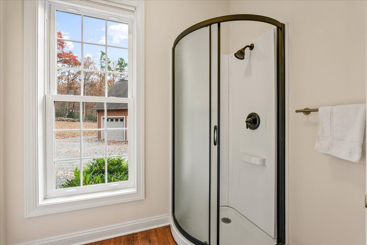 Un baño con cabina de ducha de vidrio, una toalla blanca colgada en la pared y una ventana con vistas a una casa y árboles.