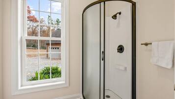 Un baño con cabina de ducha de vidrio, una toalla blanca colgada en la pared y una ventana con vistas a una casa y árboles.