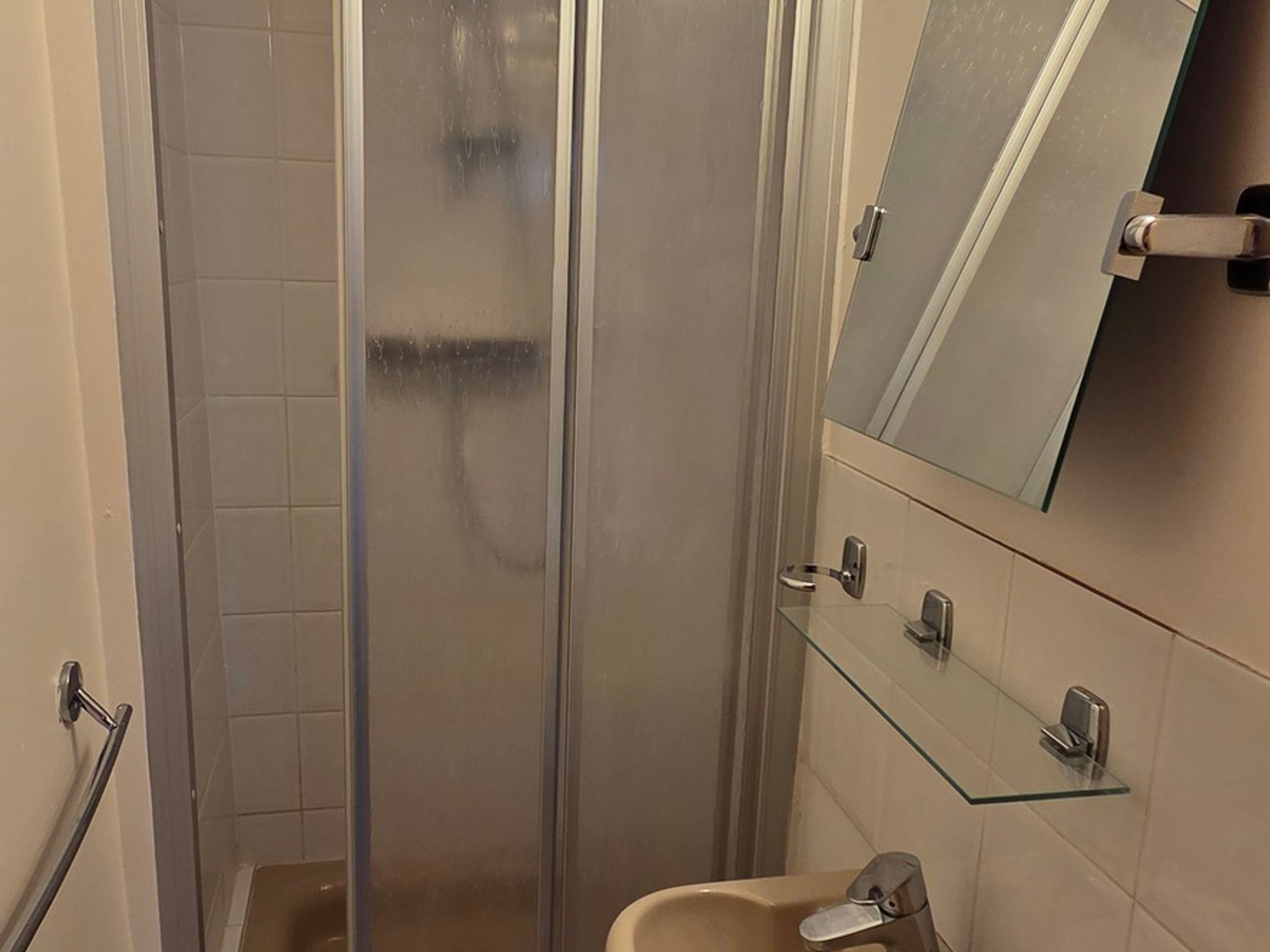 Et badeværelse med en glasbrusekabine, en glashylde med toiletartikler og et spejl over vasken.