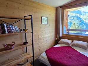 Une pièce aux murs lambrés, avec un lit, une bibliothèque et une fenêtre donnant sur les montagnes.