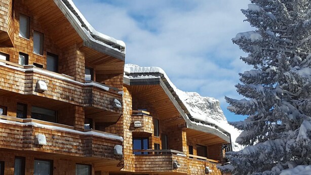 Ein mehrstöckiges Holzchalet mit Balkonen vor einer verschneiten Berglandschaft.