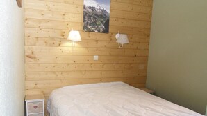 Un dormitorio con pared de madera, una cama con ropa de cama blanca, dos lámparas de noche y una mesita de noche pequeña con un cajón estampado con corazones rojos.