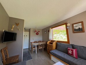 Une pièce chaleureuse avec un canapé en bois, un téléviseur, une table à manger avec des chaises et une fenêtre donnant sur un paysage verdoyant.