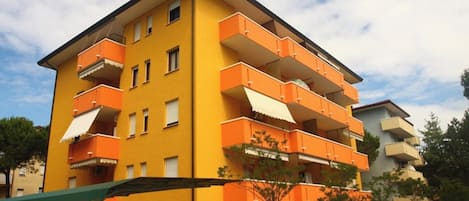 A multi-story yellow building with orange balconies, surrounded by parked cars and greenery.