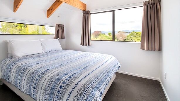 A bedroom with a large bed, wooden beams, a window with curtains, and a view of greenery. - 6 Bedroom Family Gateway in the Heart of Mapua Village (Māpua)
