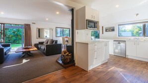 A modern living room with a sofa, a coffee table, and a fireplace. The kitchen has white cabinets, a microwave, and a dishwasher. - 6 Bedroom Family Gateway in the Heart of Mapua Village (Māpua)
