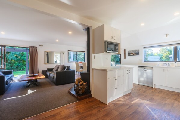A modern living room with a sofa, a coffee table, and a fireplace. The kitchen has white cabinets, a microwave, and a dishwasher. - 6 Bedroom Family Gateway in the Heart of Mapua Village (Māpua)