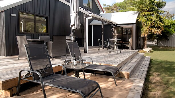A modern outdoor deck with metal chairs and a table, adjacent to a dark-colored building with large windows and a covered patio area. - 6 Bedroom Family Gateway in the Heart of Mapua Village (Māpua)