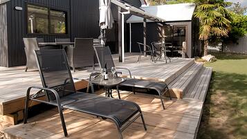 Una terraza moderna al aire libre con sillas de metal y una mesa, contigua a un edificio de color oscuro con grandes ventanales y un porche cubierto.