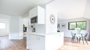 A modern kitchen with white cabinetry, a microwave, and a dining area with a table and chairs. - 6 Bedroom Family Gateway in the Heart of Mapua Village (Māpua)
