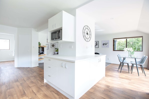 A modern kitchen with white cabinetry, a microwave, and a dining area with a table and chairs. - 6 Bedroom Family Gateway in the Heart of Mapua Village (Māpua)