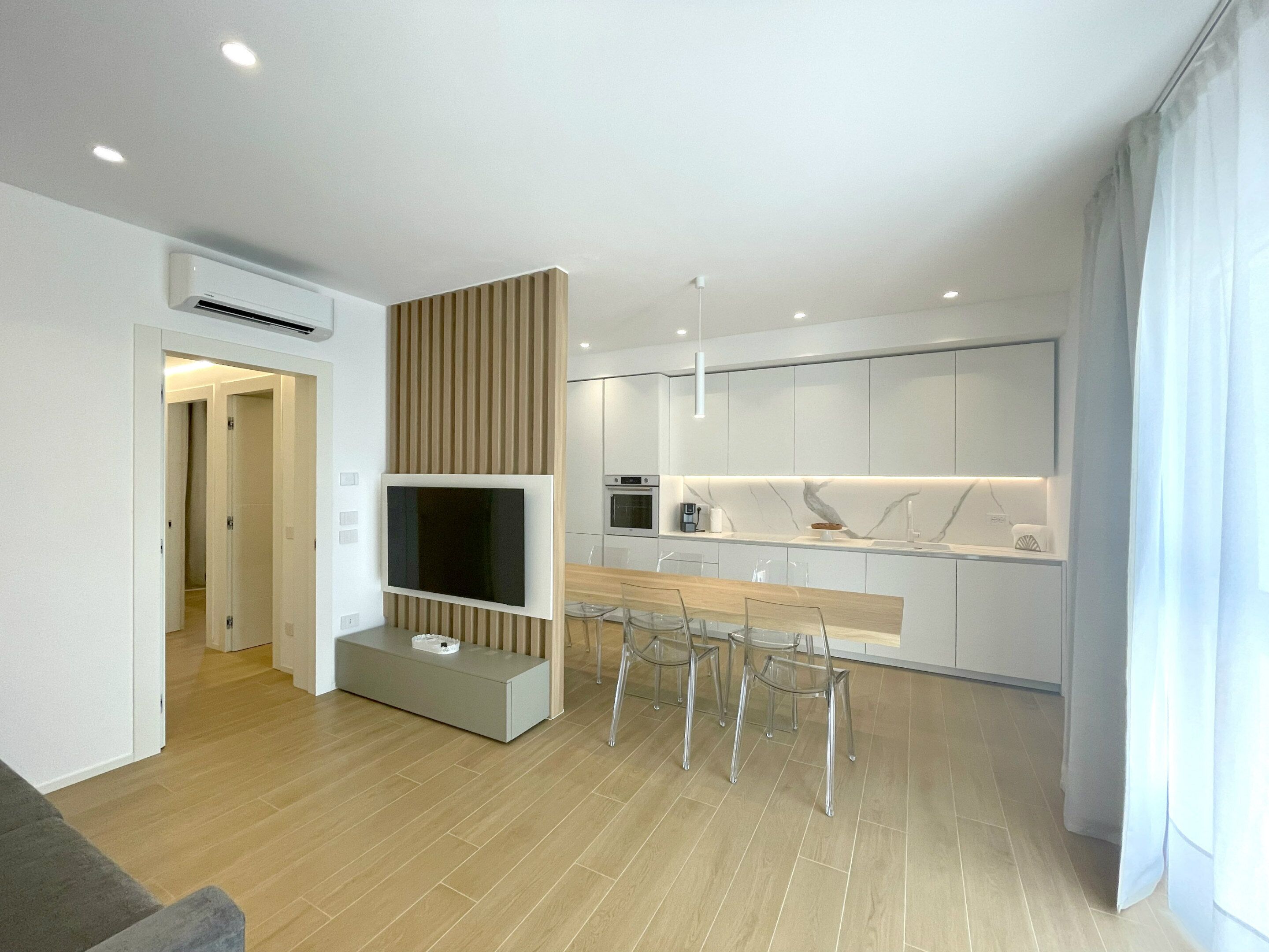 Eine moderne Küche mit Einbauofen, ein Essbereich mit Holztisch und transparenten Stühlen sowie ein an der Wand befestigter Fernseher.