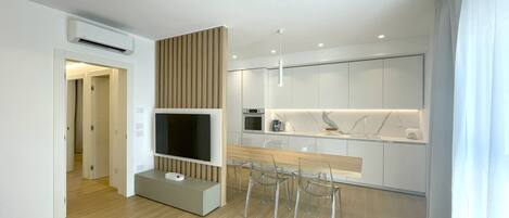 Eine moderne Küche mit Einbauofen, ein Essbereich mit Holztisch und transparenten Stühlen sowie ein an der Wand befestigter Fernseher.