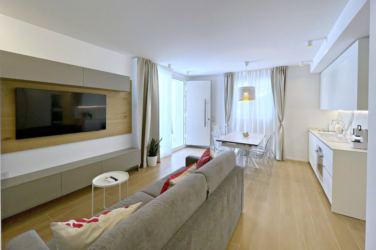 A modern living room with a flat-screen TV, a grey sofa, a dining table with chairs, and a kitchen area with a white countertop and cabinets.