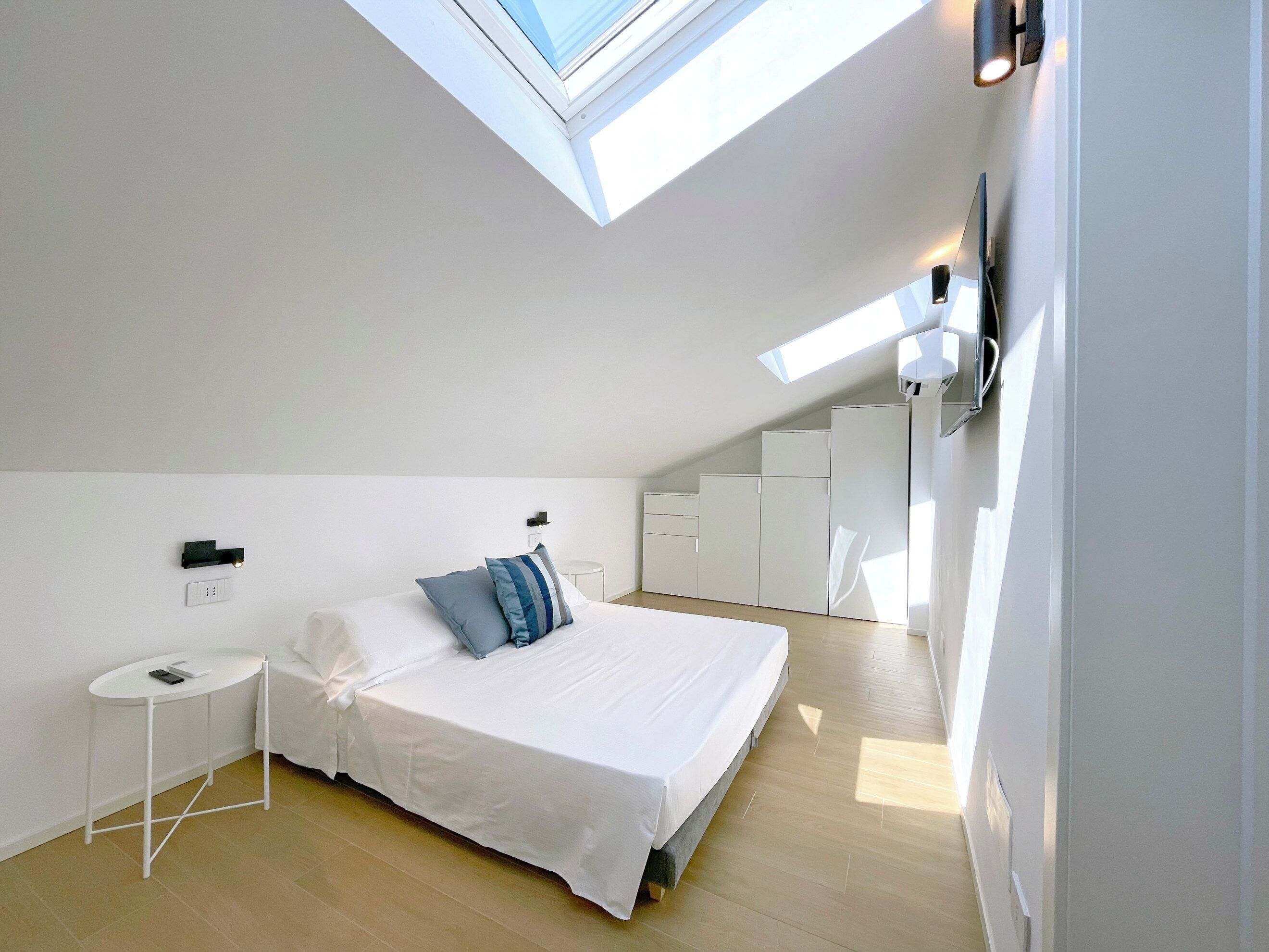 A modern bedroom with a skylight, a bed with pillows, a bedside table, and built-in wardrobes.