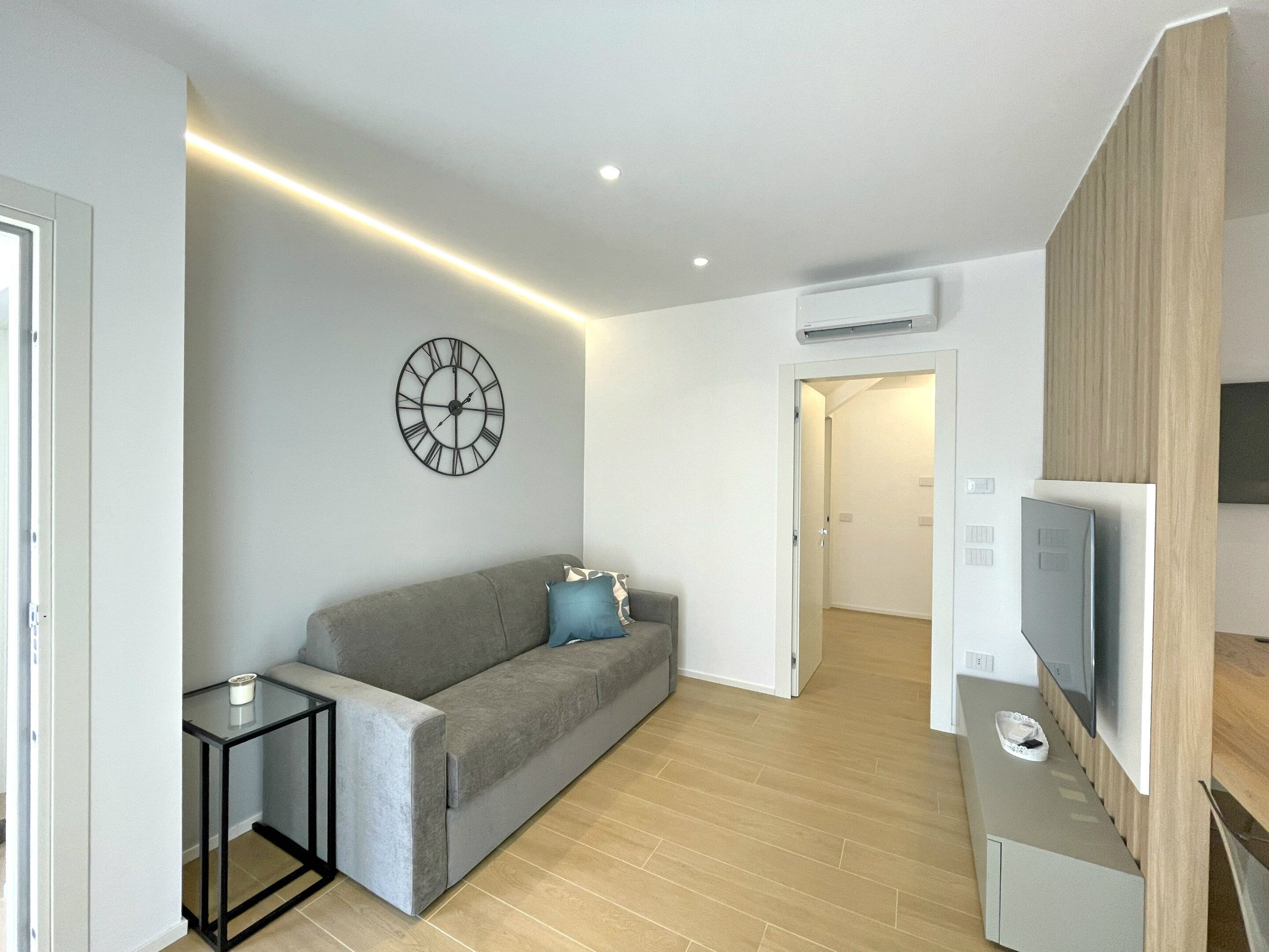 Een moderne woonkamer met een grijze bank, een klein bijzettafeltje, een wandklok en een flatscreen televisie bevestigd aan een houten paneel.