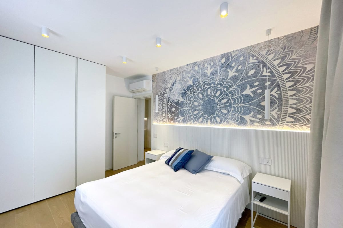 A modern bedroom with a large bed, a wardrobe, a nightstand, and a decorative wall piece.