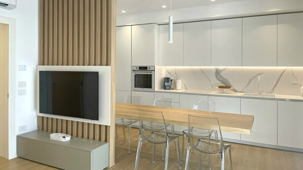 Una cocina moderna con horno empotrado, una zona de comedor con mesa de madera y sillas transparentes, y un televisor montado en la pared.