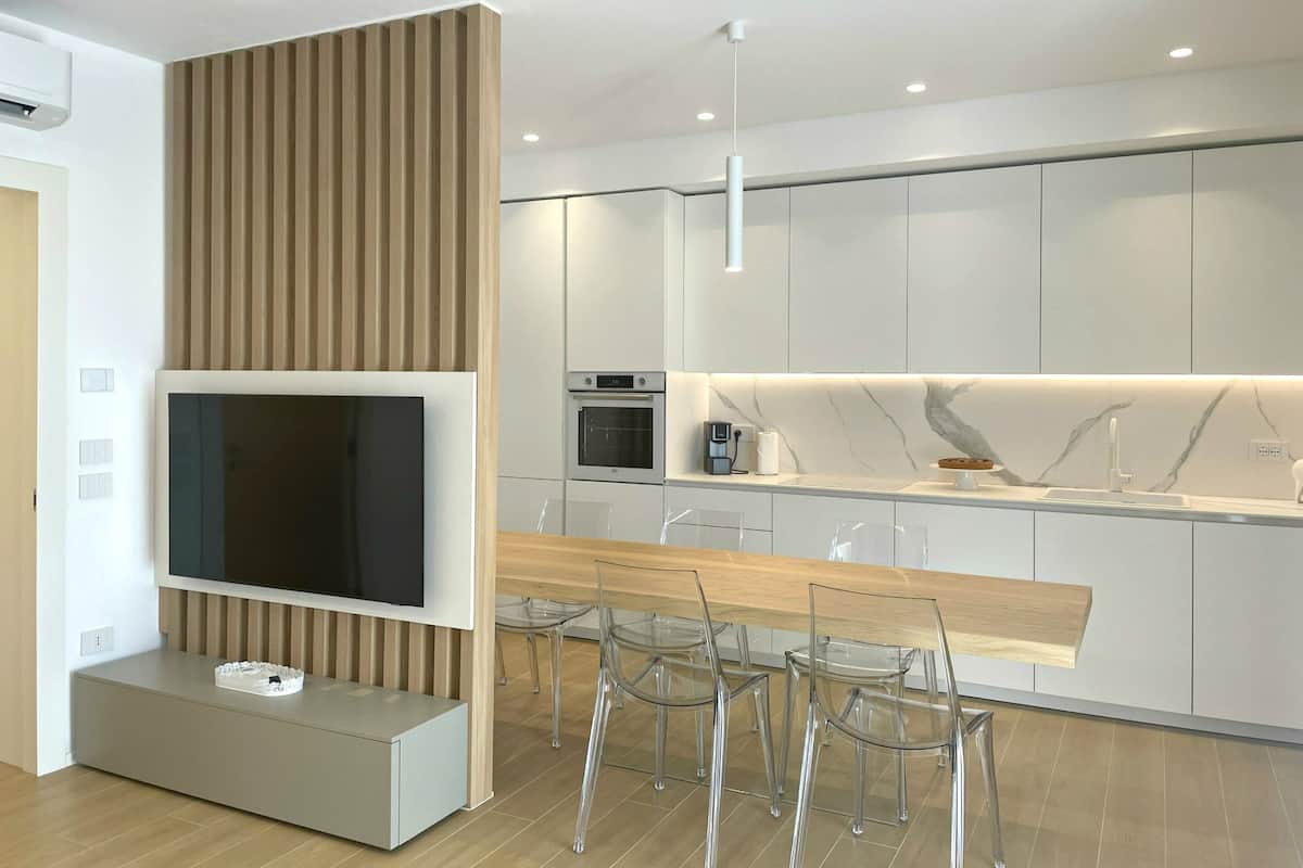 A modern kitchen with a built-in oven, a dining area with a wooden table and clear chairs, and a TV mounted on a wall.