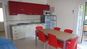 A compact kitchen with red cabinets, a microwave, and a refrigerator. There is a dining table with red chairs and a blue sofa in the foreground. - Charming Villa 500mt From the Beach (San Michele al Tagliamento)
