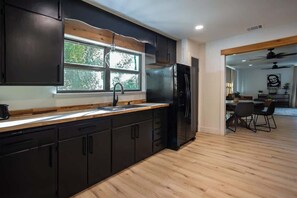 A modern kitchen with dark cabinetry, a black refrigerator, a window with a view, and a dining area with chairs.