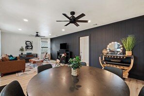 A modern living room with a dark wall, a ceiling fan, a round dining table, and a television. - Das Green Dor - Sleeps 9, Hot Tub & Fire Pit (Fredericksburg)