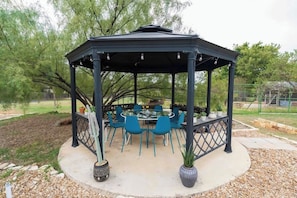 A gazebo with a glass top table and blue chairs, surrounded by a gravel area and trees.