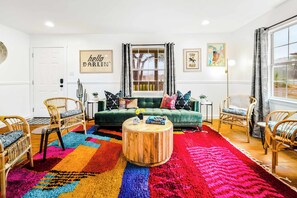 A living room with a green sofa, wicker chairs, a colorful rug, and a wooden coffee table. - Howdy Haus - Pool, Hot Tub, Game Room & Glam Room (Fredericksburg)