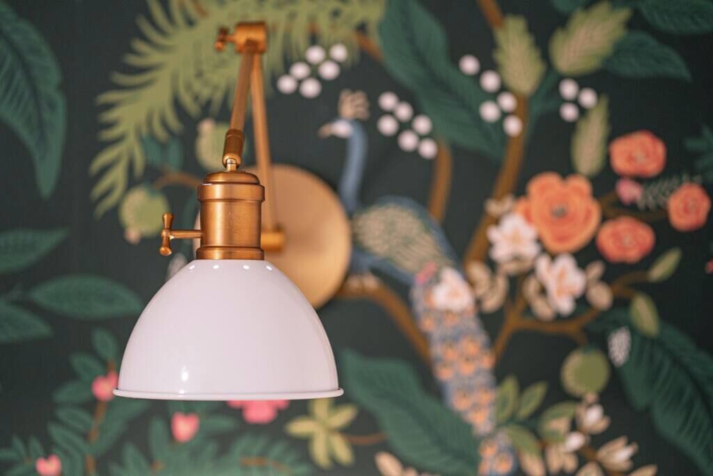 Eine weiße Lampe mit Messingsockel vor floralem Wandpapier.