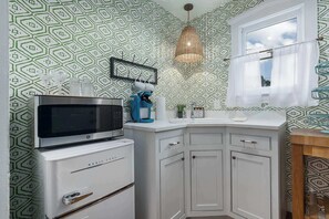Una cocina con microondas, refrigerador, gabinetes blancos, una lámpara colgante y una ventana con cortinas.