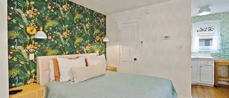 A bedroom with a bed, a nightstand, a lamp, a door, and a view of a bathroom.