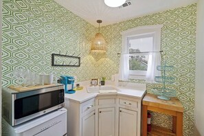 Cocina compacta con microondas, lavamanos y armarios, que presenta papel tapiz de estampado geométrico y una lámpara colgante.