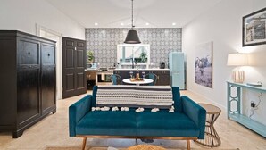 A modern living room with a blue sofa, a dining table with chairs, a kitchen in the background, and a pendant light. - Stonewall Bungalow 3 - Sleeps 4 (Stonewall)