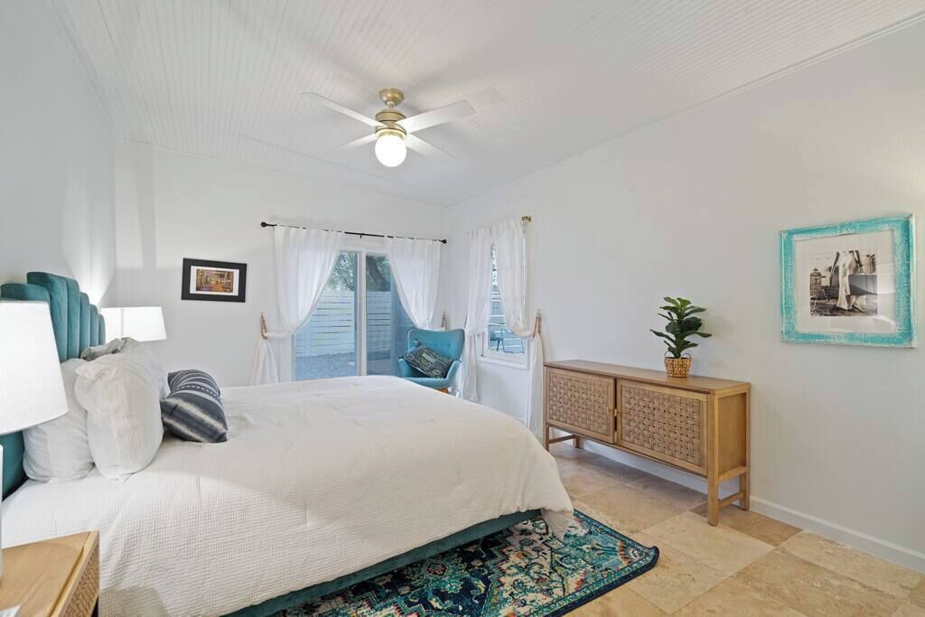 A bedroom with a bed, a ceiling fan, a window with curtains, a bedside table, a nightstand, a dresser, and a framed picture on the wall.