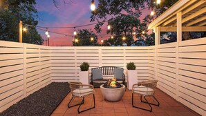 A patio with a fire pit, two wicker chairs, a bench, and string lights against a sunset sky. - Stonewall Bungalow 3 - Sleeps 4 (Stonewall)