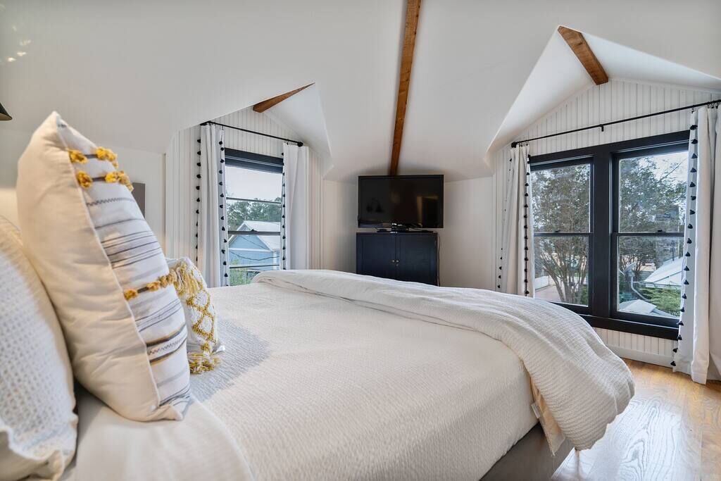 A bedroom with a large bed, a TV, and two windows with curtains.