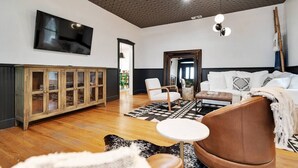 A spacious living room with a wooden cabinet, a television mounted on the wall, a sofa, a coffee table, and a patterned rug. - Stonewall Main Haus - Sleeps 6 (Stonewall)